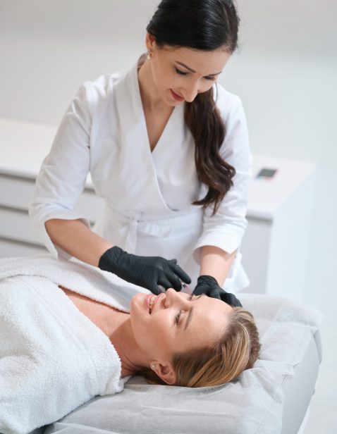 Brunette esthetician makes beauty injections to a blonde woman, female in a bathrobe lies on a cosmetology couch
